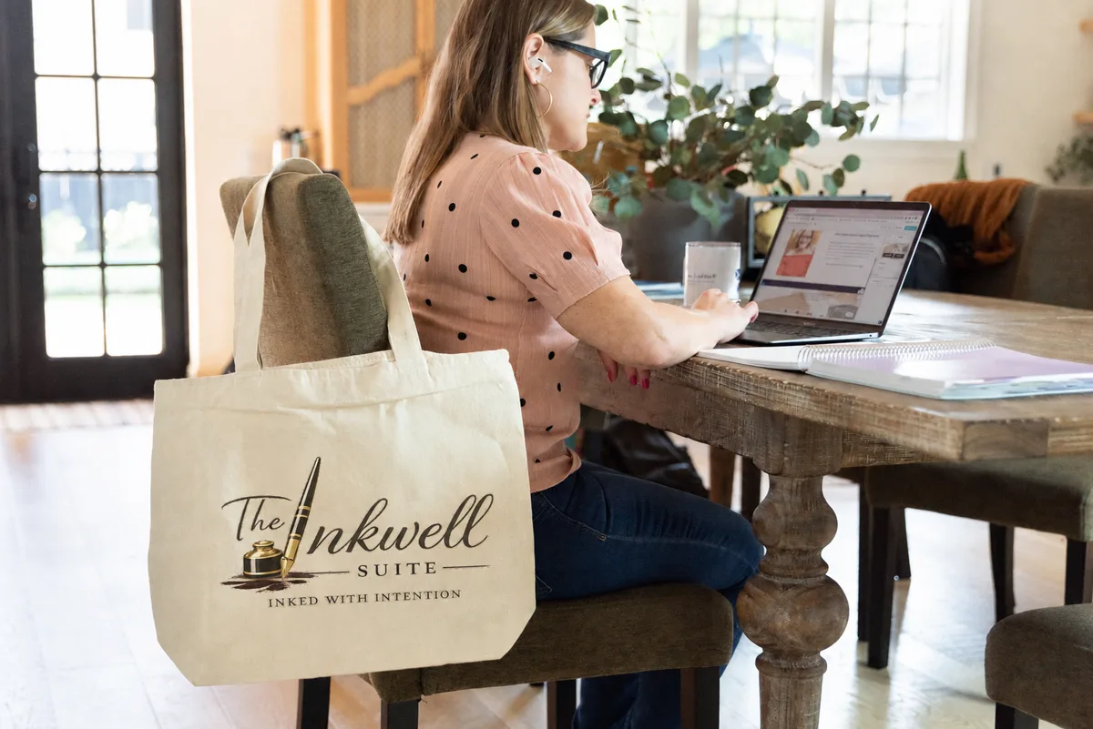 A woman working at a laptop at a table, with a canvas tote bag reading “The Inkwell Suite, Inked With Intention” hanging from her chair